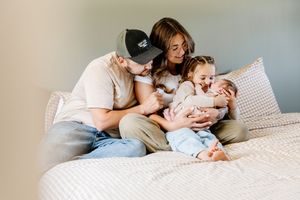 Family of four sitting on bed, parents lovingly gazing at newborn baby while toddler smiles nearby.
