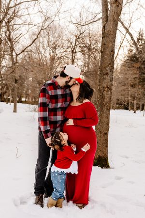 Family kissing in snowy park, pregnant mom in red, dad in plaid, toddler between them.