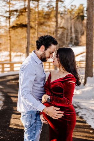 Couple sharing a kiss outdoors in winter, woman in red velvet dress with baby bump.