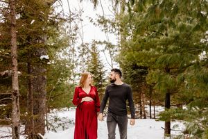Pregnant woman in red dress and man walking hand in hand through snowy pine forest.