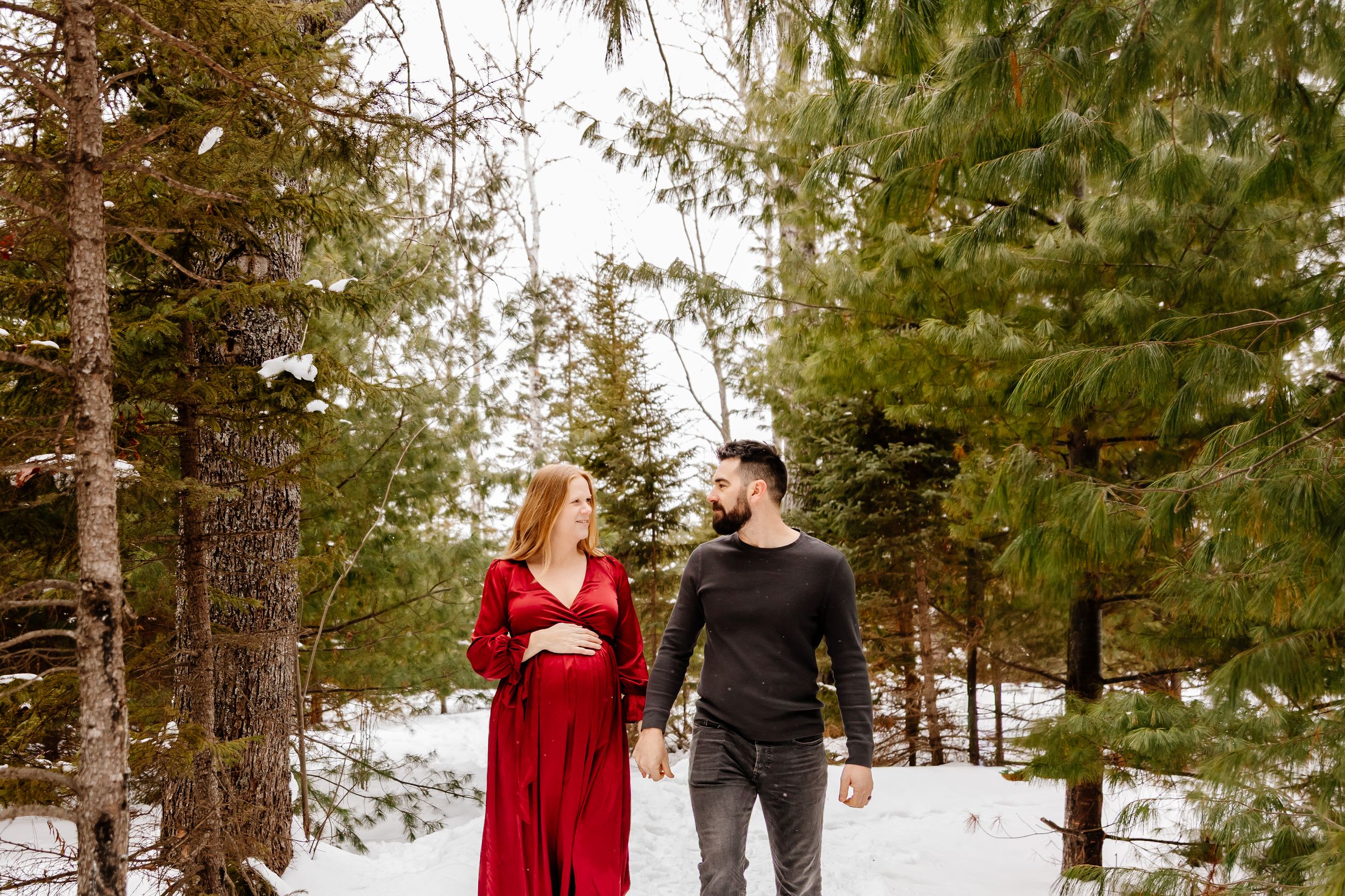 Pregnant woman in red dress and man walking hand in hand through snowy pine forest.