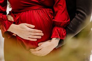 Close-up of couple's hands cradling pregnant belly in red satin maternity dress outdoors.