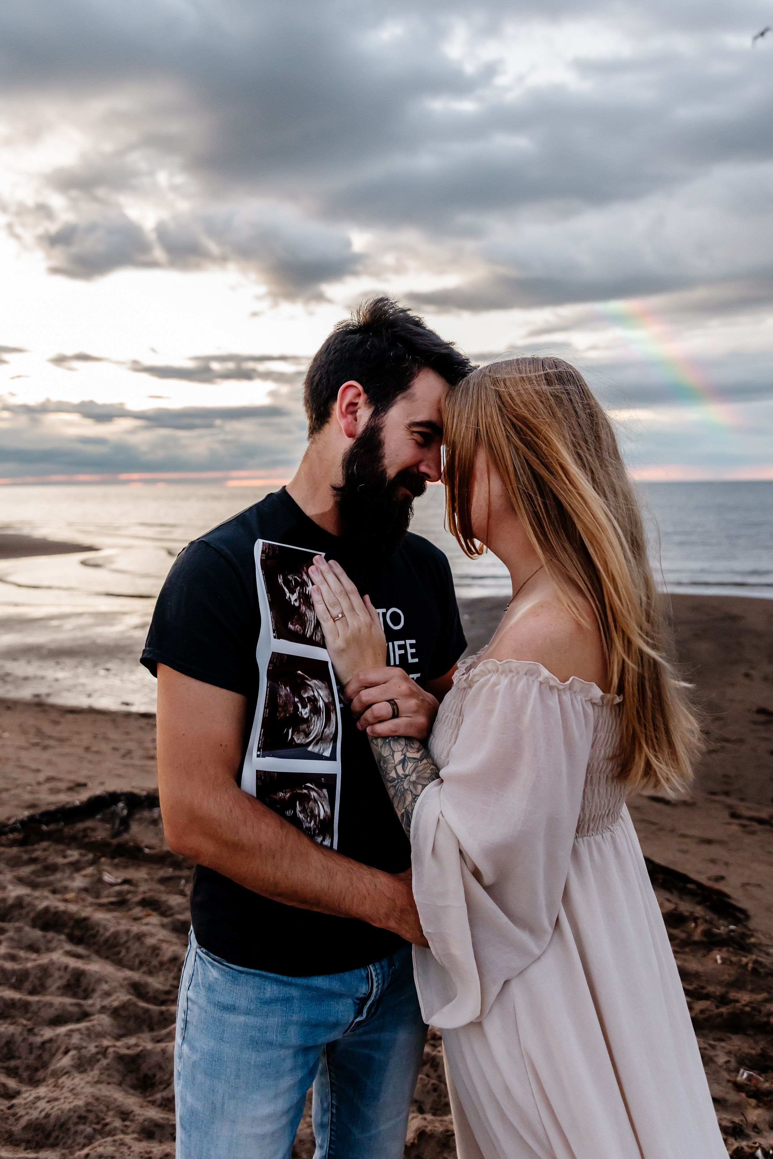 Couple touching foreheads on a beach at sunset with a rainbow in the cloudy sky.
