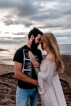 Couple touching foreheads on a beach at sunset with a rainbow in the cloudy sky.