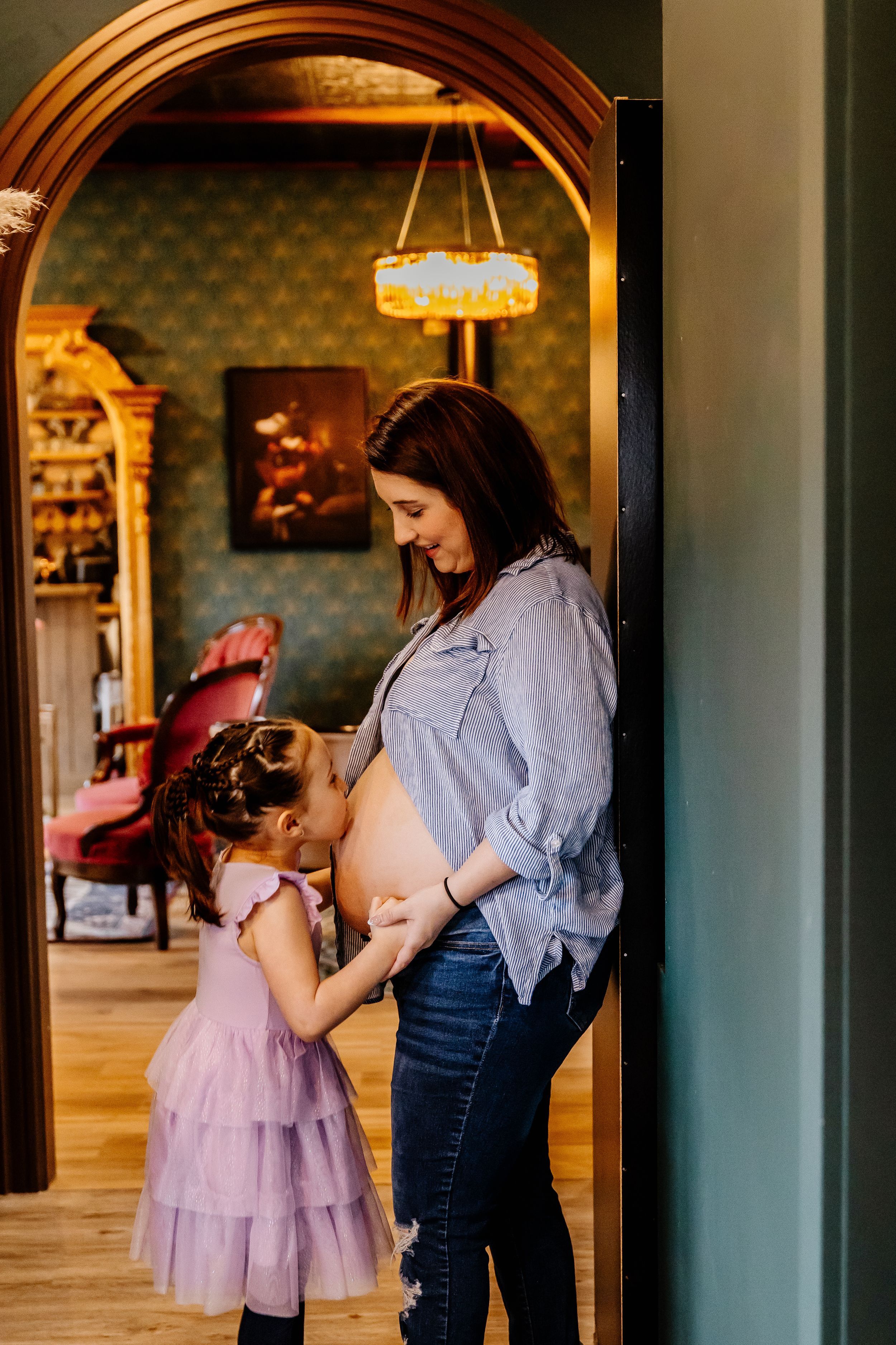 Young girl in purple dress kissing her mother's pregnant belly in an elegant home.