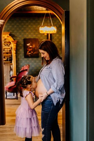 Young girl in purple dress kissing her mother's pregnant belly in an elegant home.