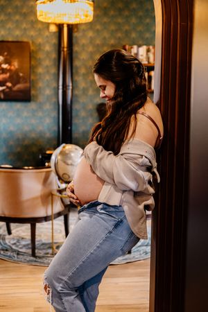 Pregnant woman in jeans cradling her baby bump while leaning against a doorway indoors.