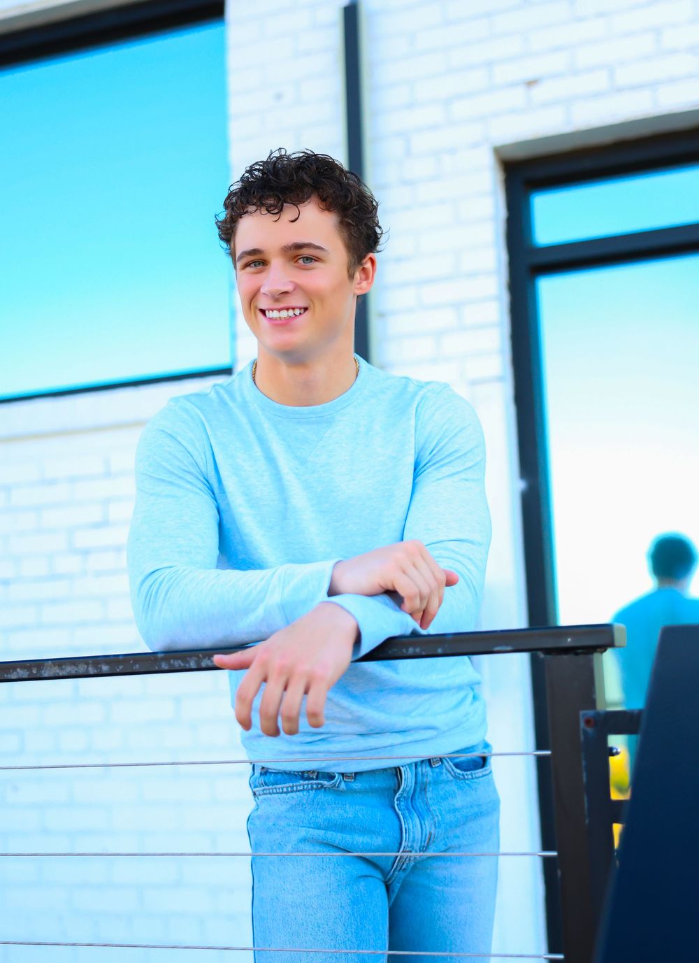 Person in light blue sweater and jeans leaning against a railing with a white brick wall background.