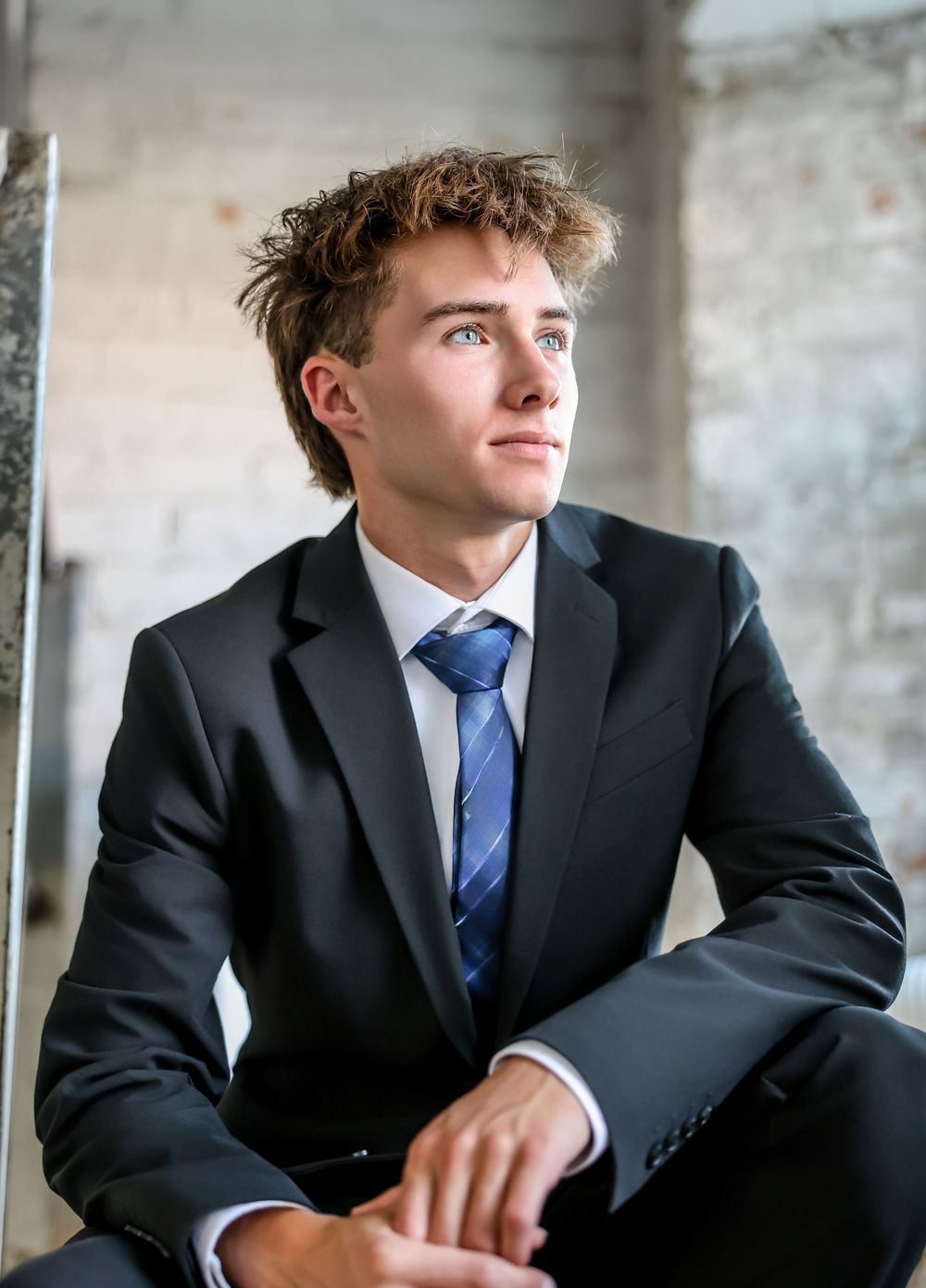 Young professional in dark suit and blue tie sits thoughtfully in an industrial setting with multiple poses.