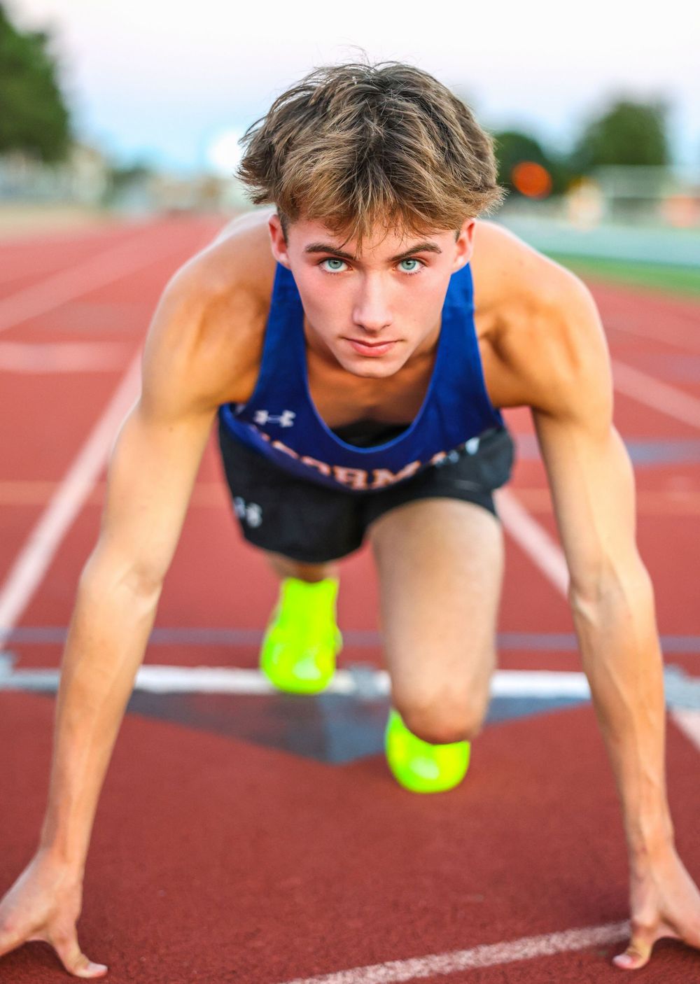 Athlete in blue uniform crouches in starting position on red running track wearing neon yellow shoes.