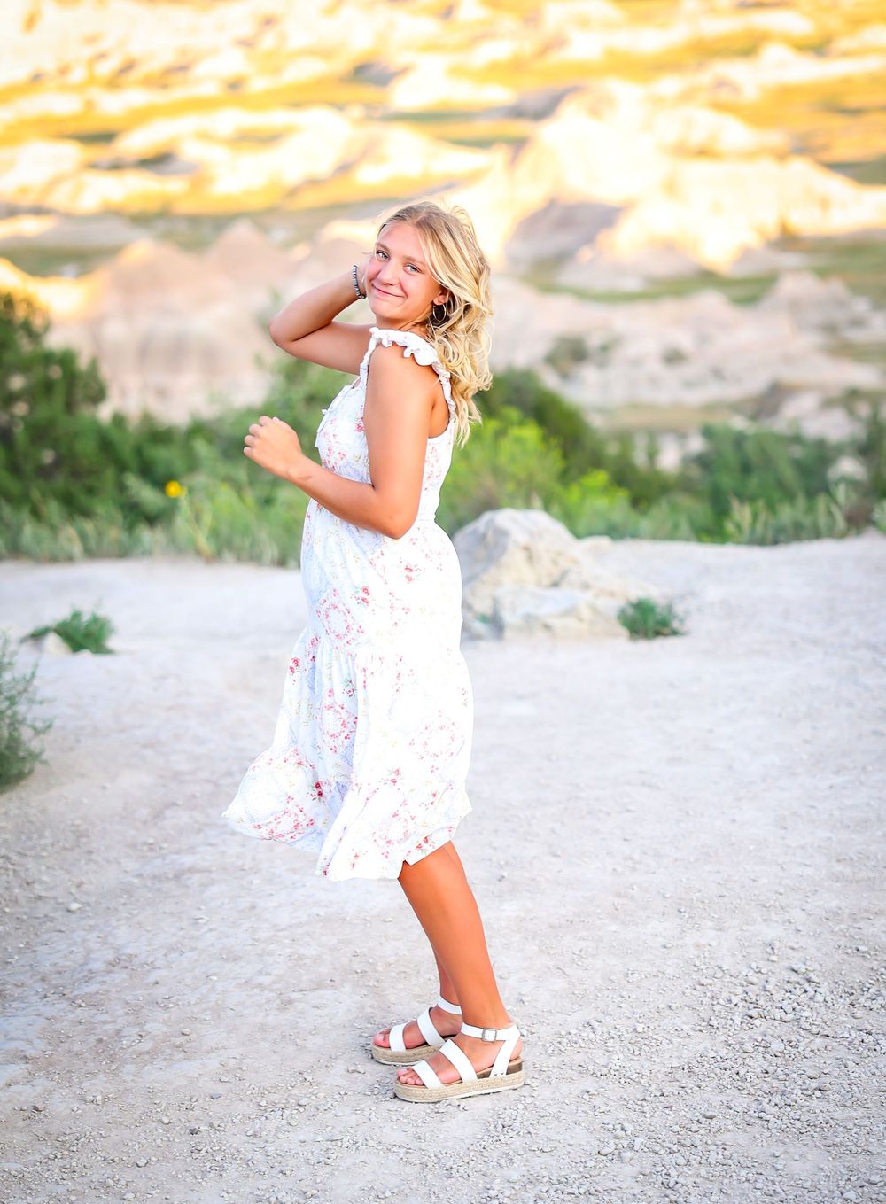 Playful poses in white floral dress against sandy outdoor setting.