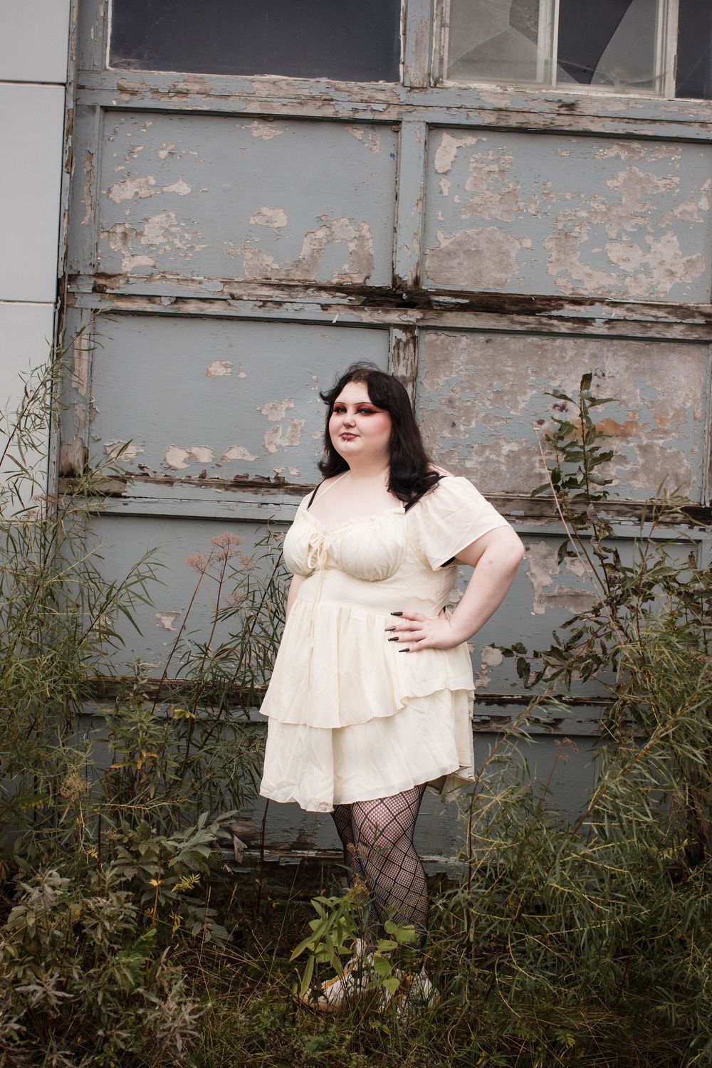 A person in a cream-colored tiered dress and patterned tights poses against a weathered industrial wall with overgrown foliage.