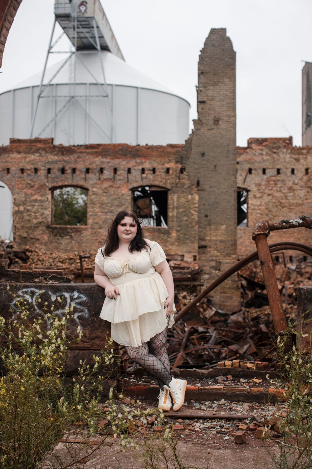 A person in a cream-colored dress poses on rustic stone steps against ruins and an industrial structure in the background.