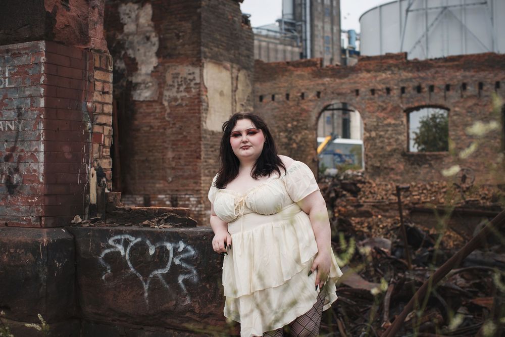 A person in a cream-colored vintage dress poses against weathered brick ruins with industrial buildings in the background.