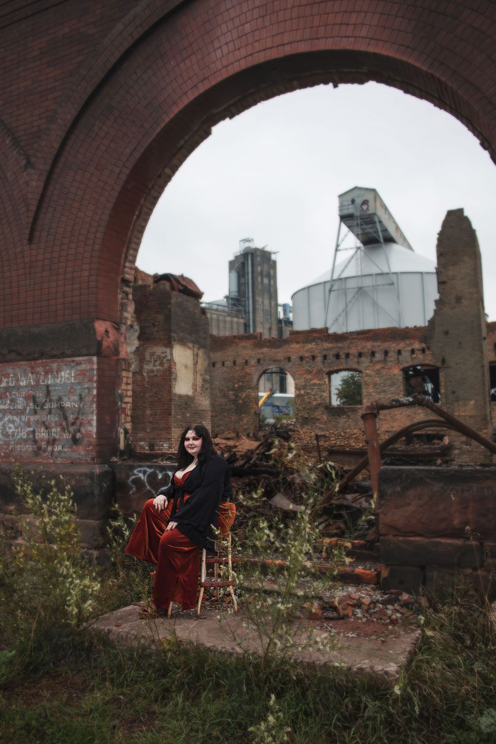 A person in dark clothing poses in an abandoned brick archway surrounded by urban decay and graffiti.