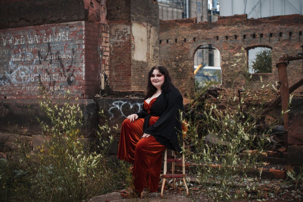 A woman in a flowing red dress poses dramatically against rustic brick walls in an urban outdoor setting.