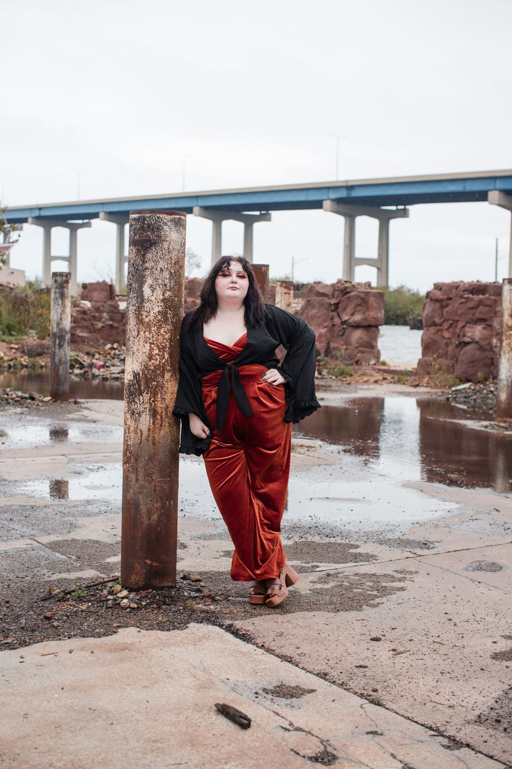 A person in a flowing red velvet dress poses dramatically against rustic wooden posts in an industrial setting.