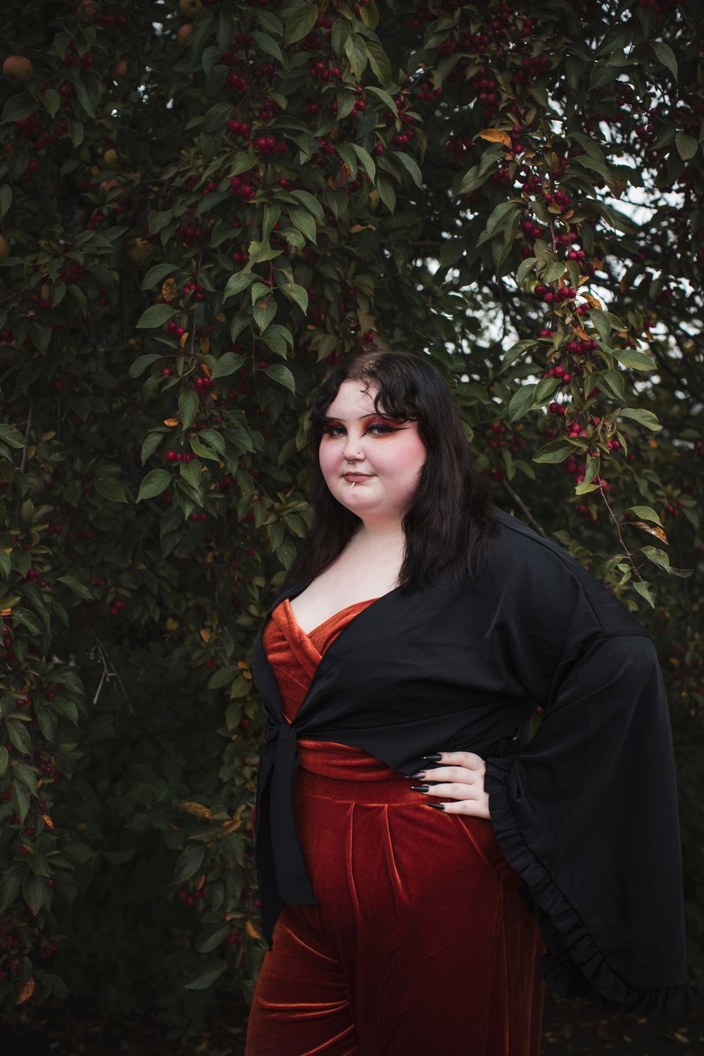 A figure in a red velvet dress and black cardigan poses against a backdrop of dark foliage with red berries.