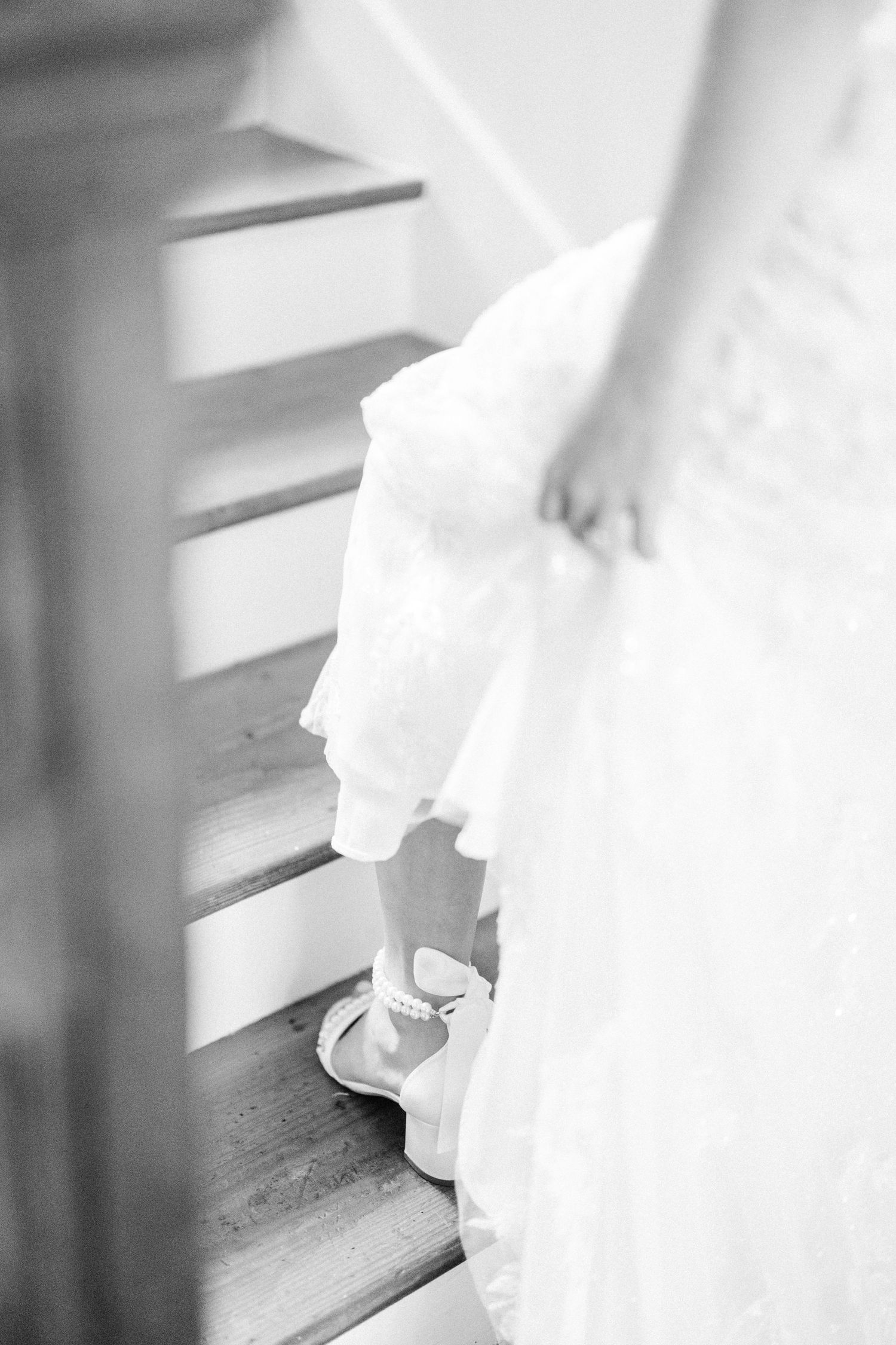 A black and white photograph shows a bride's white dress and elegant shoe on wooden stairs.