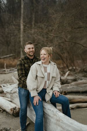 A couple shares a joyful moment together while sitting on driftwood at a beach during golden hour. Engagment Photography - Hudson Valley