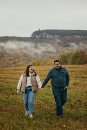 A couple walks together through a misty autumn field with cliffs and fall foliage in the background. -Engagment Photography - Hudson Valley