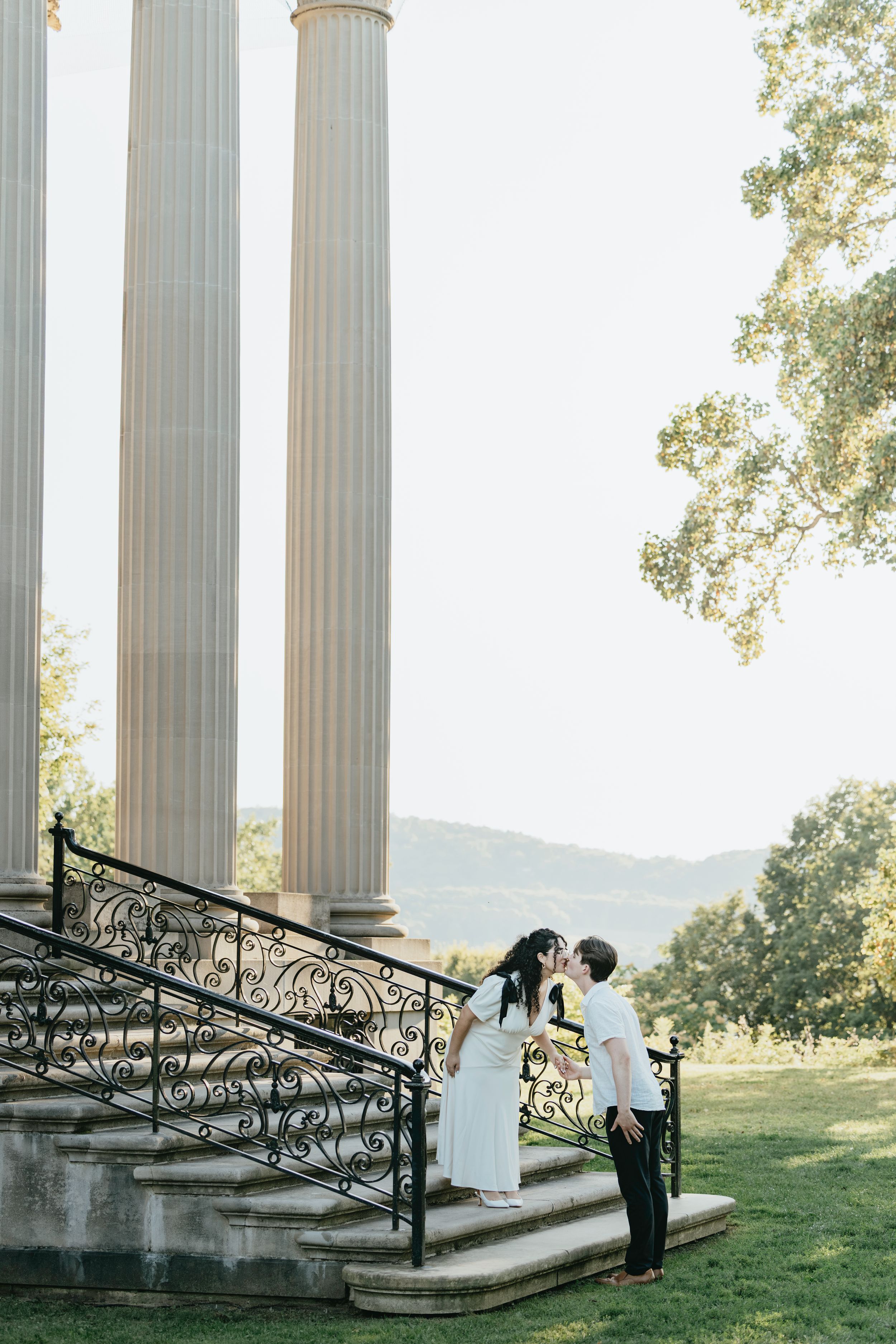Elegant couple shares a romantic moment on white marble steps surrounded by classical columns in an outdoor setting.