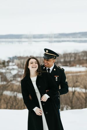 A couple shares a joyful moment in the snow, with one wearing a military dress uniform against a winter landscape backdrop.