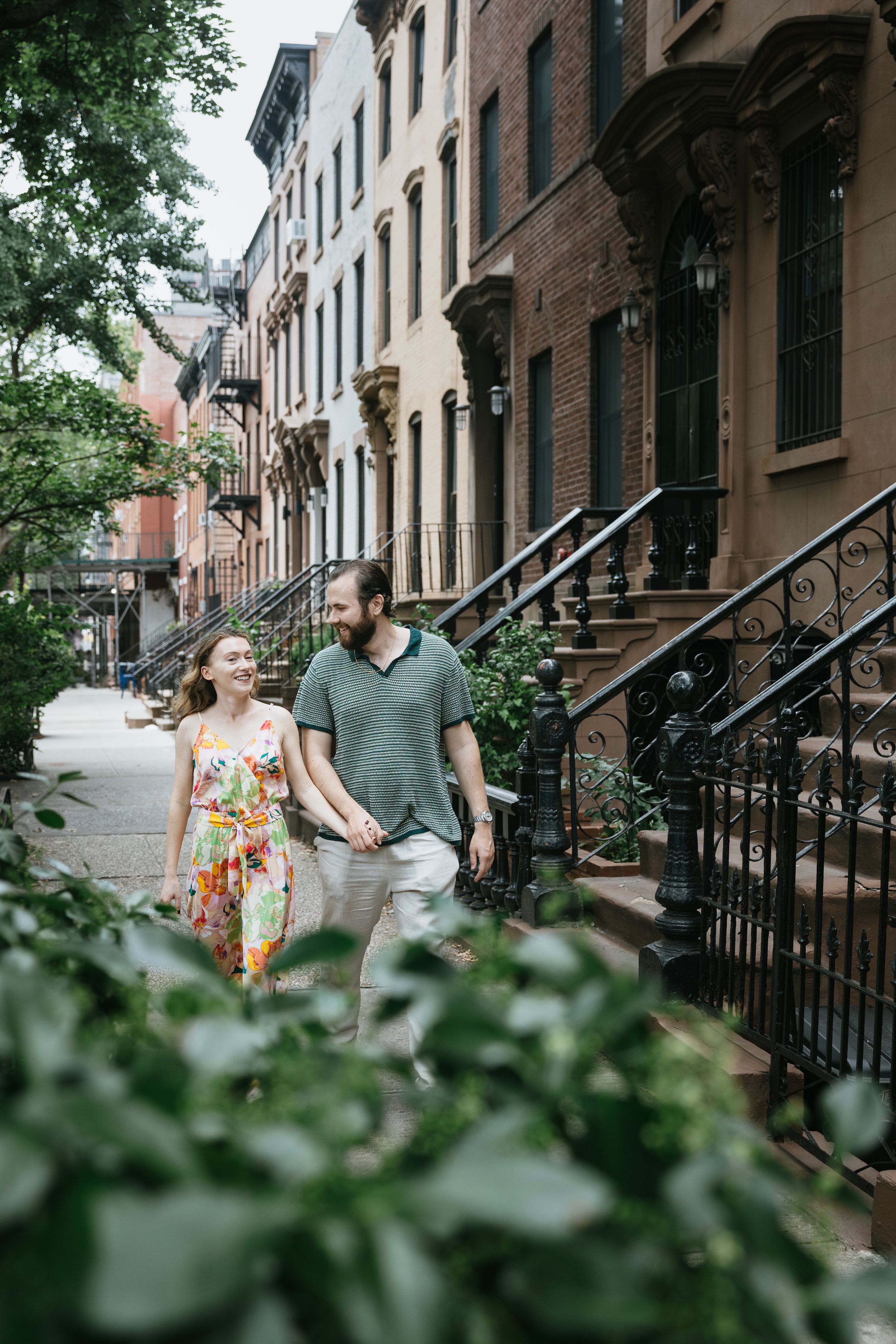 Couple walking hand in hand down a tree-lined brownstone street in a historic neighborhood on a summer day.
