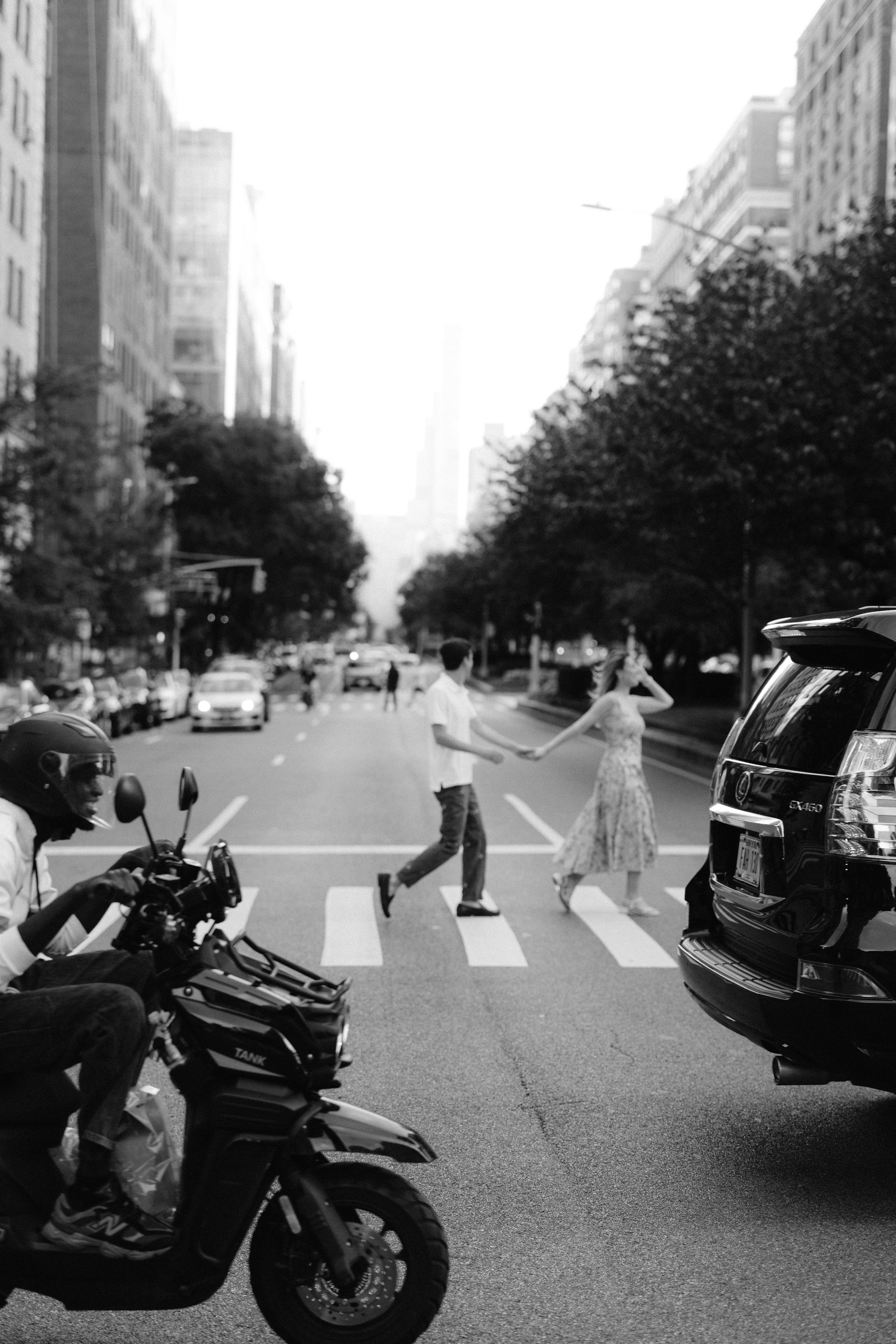 Black and white panoramic street scene showing a motorcycle and cars on an urban street with crosswalks and trees.