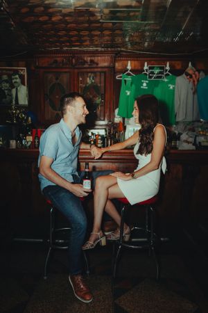 Two people enjoy drinks and conversation while sitting at a retro-style bar with sports jerseys visible in the background.Engagement Photogr