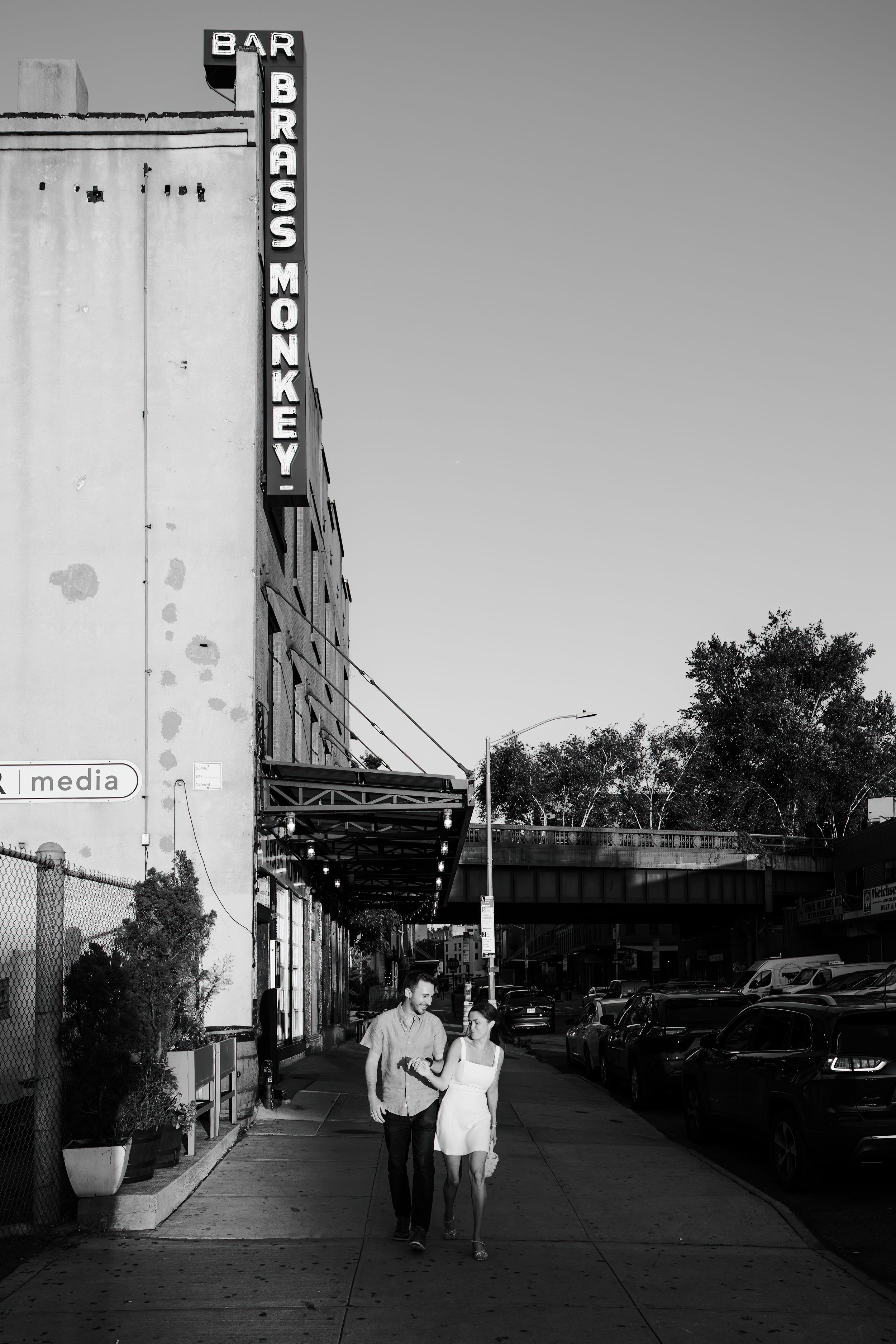 A couple walks down a city sidewalk beneath the vintage Brass Monkey bar sign in black and white.
