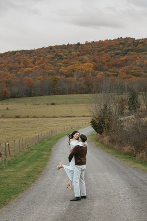 A couple embraces on a country road with autumn foliage covered hills in the background on an overcast day.