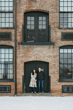 A couple stands in front of a rustic brick building with industrial windows during winter.
