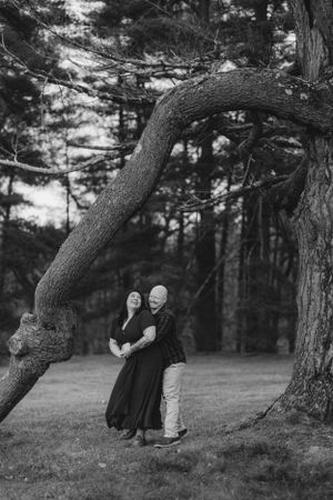 A couple embraces under a curved tree branch in a moody black and white forest setting. -Engagment Photo - Hudson Valley - Vanderbilt