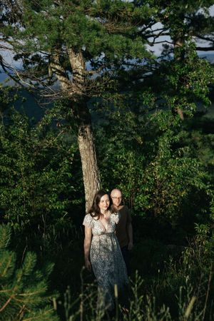 A couple stands together in a natural setting with tall pine trees and lush greenery on a sunny afternoon.