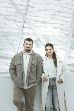 A couple in casual winter attire poses together against a white industrial background.