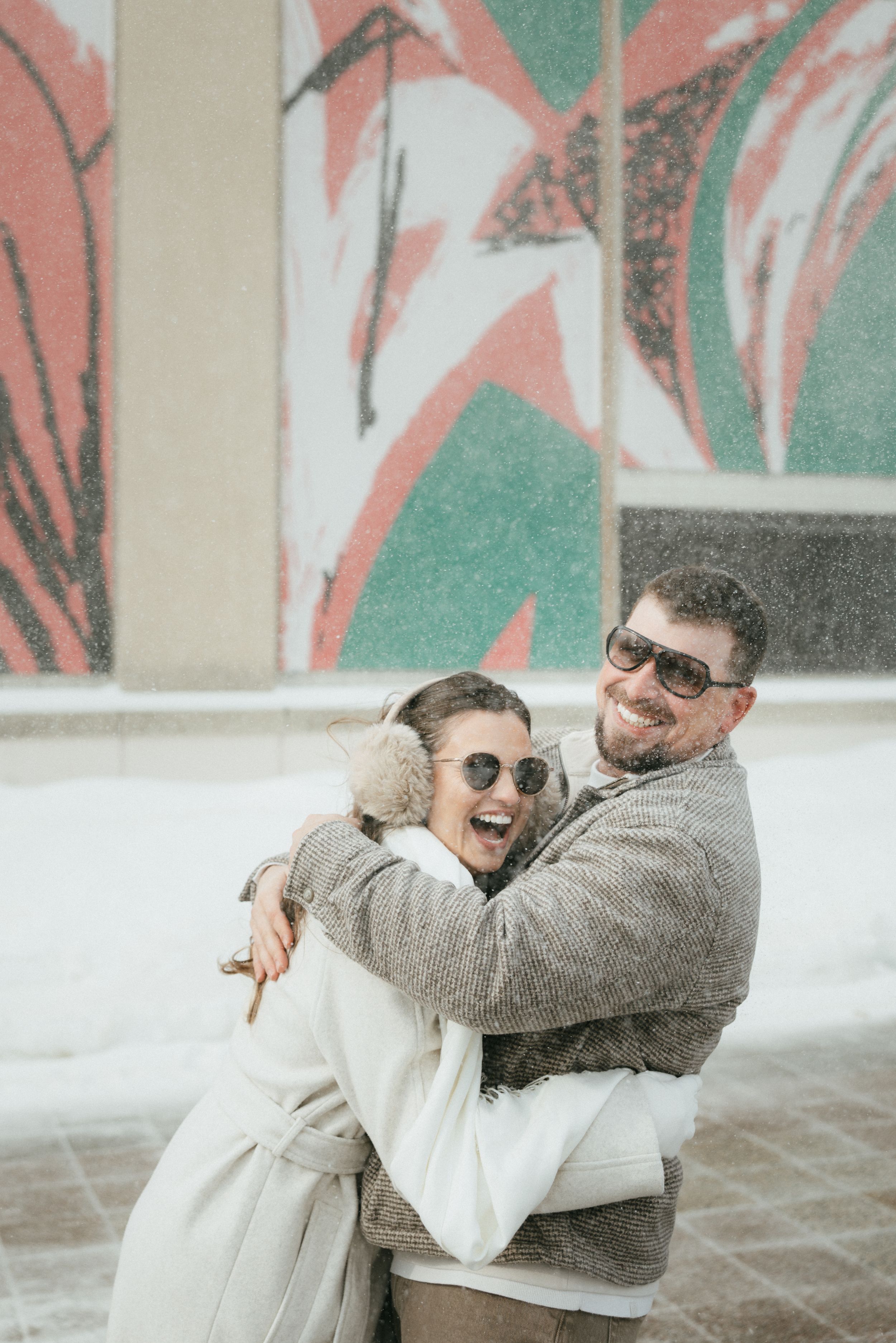 Two people embracing and laughing together against a wall with green and pink abstract artwork.