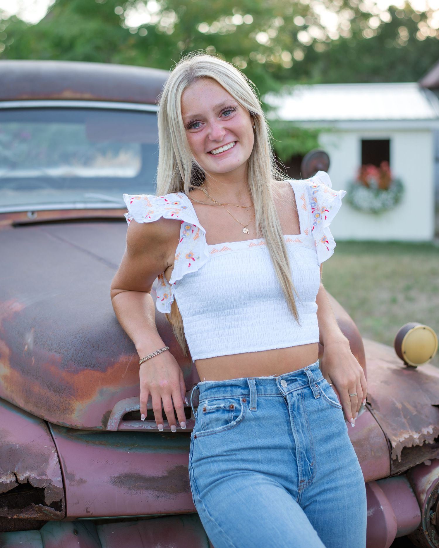 A young woman in a white crop top and blue jeans poses against a rustic vintage car outdoors.