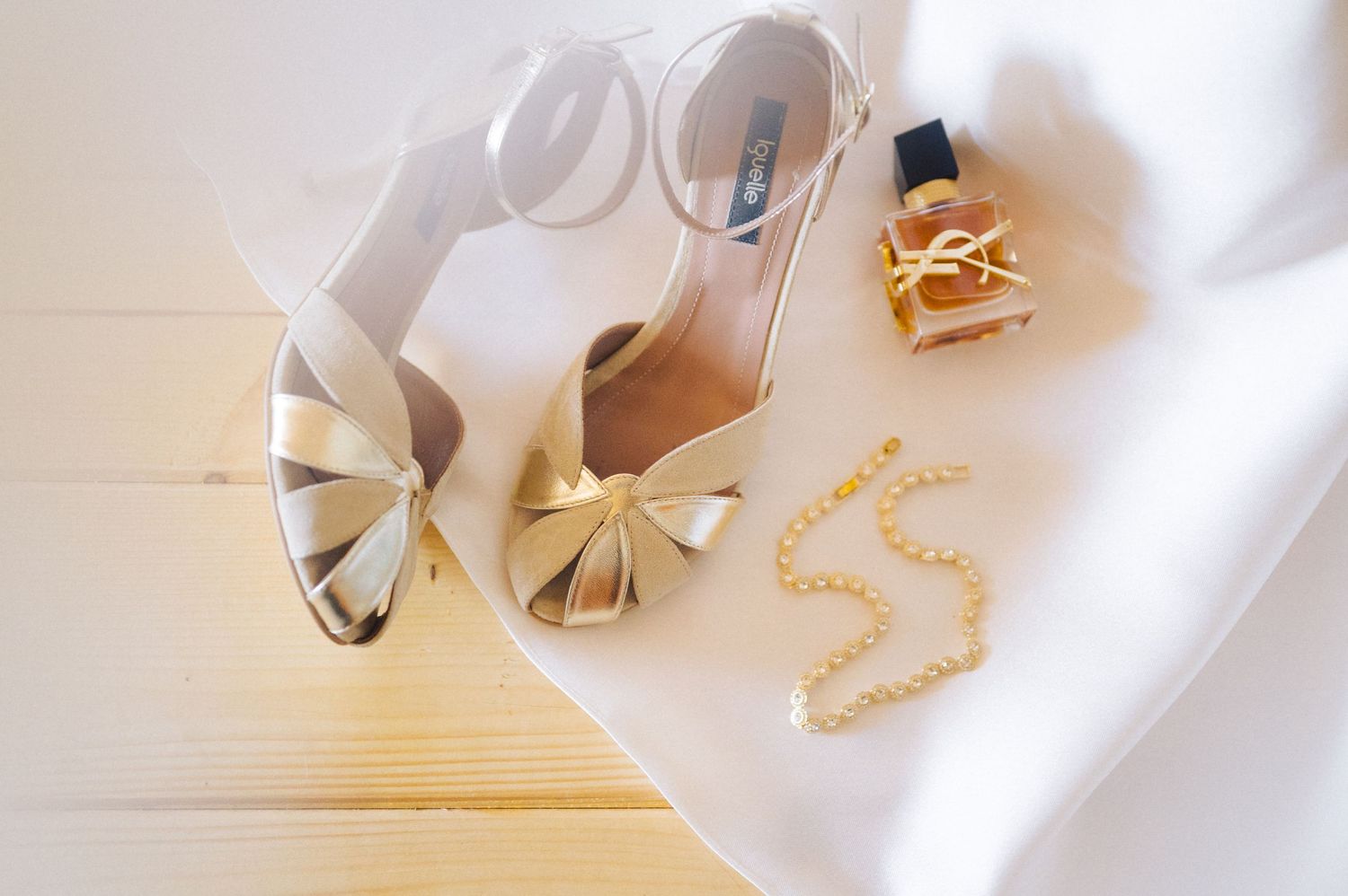 Elegant white bridal sandals with bow details displayed alongside delicate gold jewelry on white fabric.