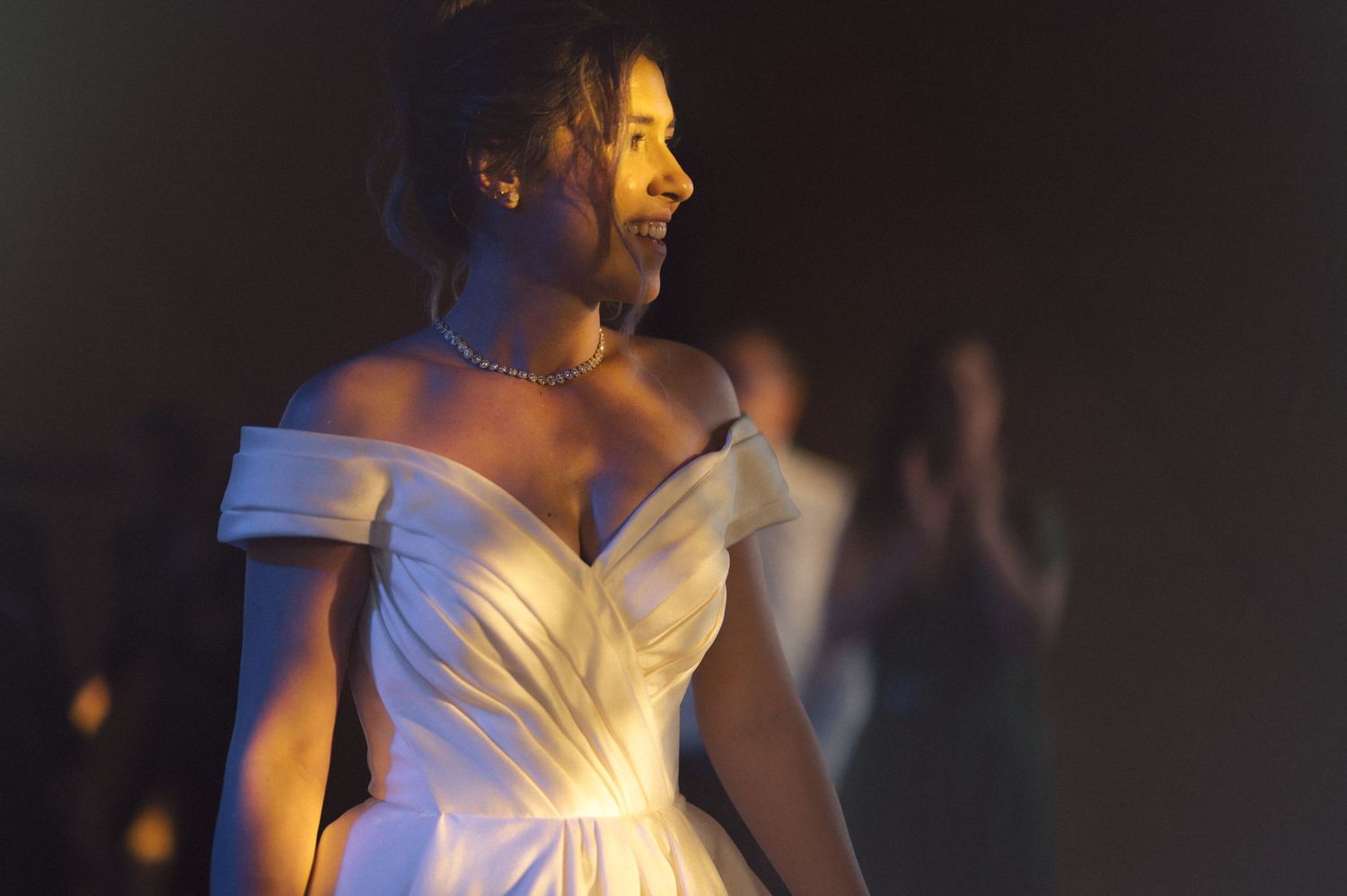 A person in an elegant off-shoulder white wedding gown stands illuminated in dramatic lighting.