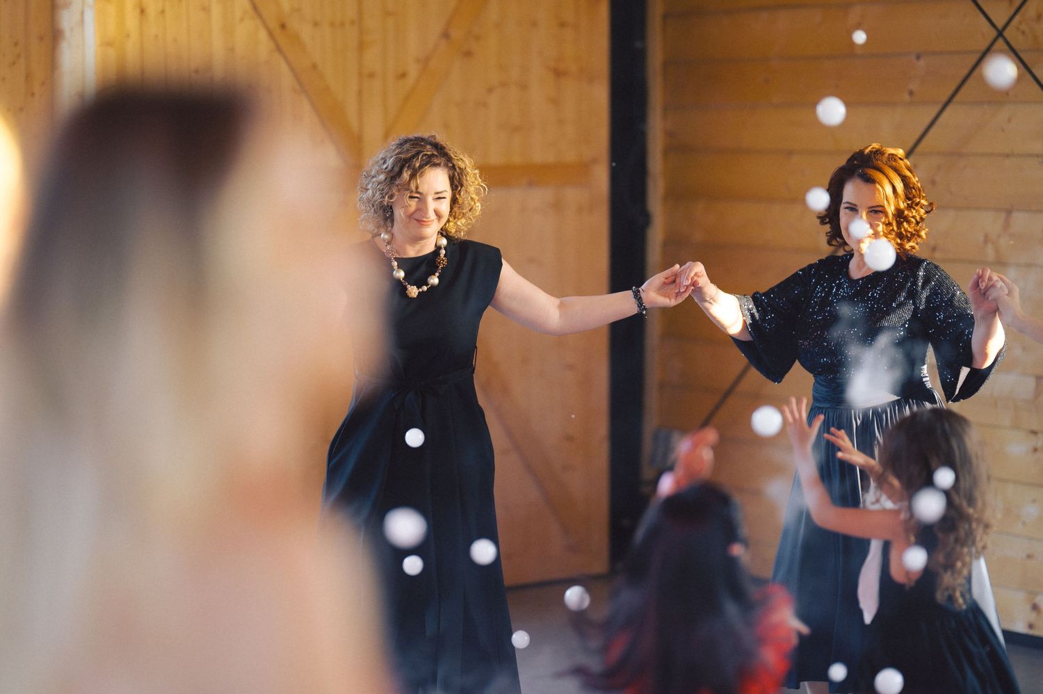 Wedding guests dancing joyfully together at a wooden-beamed venue with floating bubbles in the air.