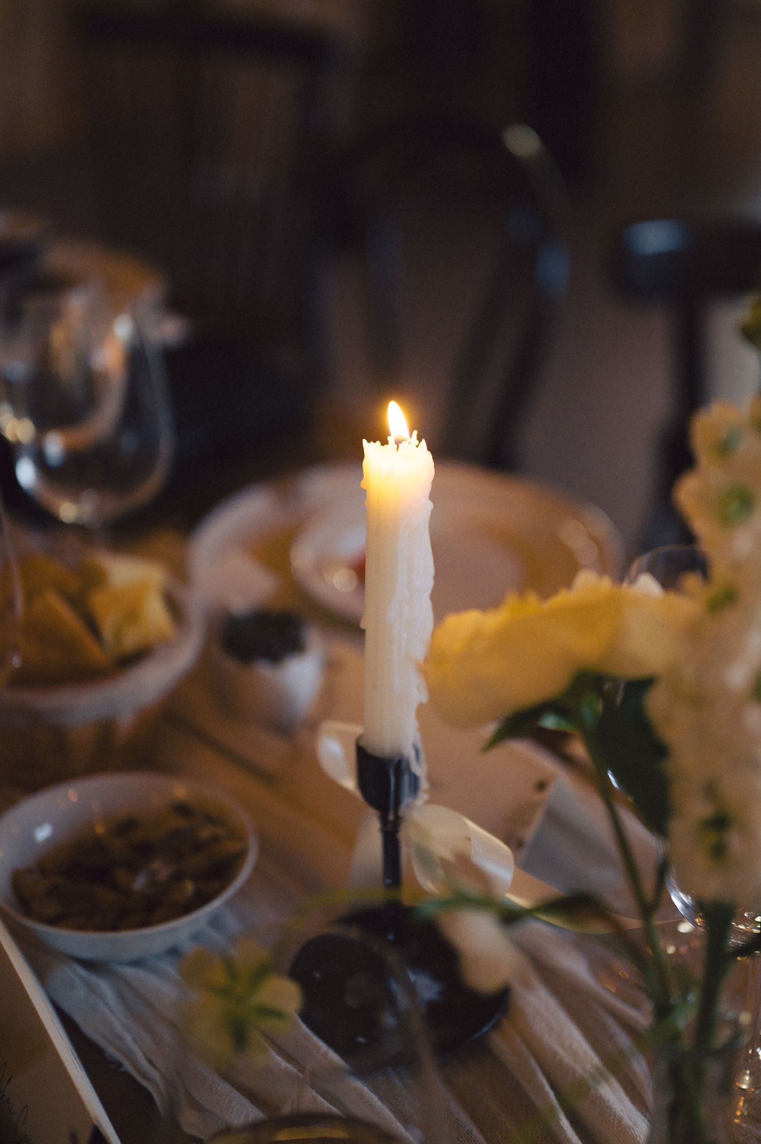 A lit white candle glows on a rustic wooden table with blurred flowers and table settings in the background.