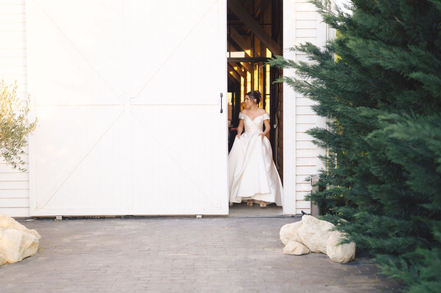 A white wedding dress peeks through an open doorway framed by evergreen trees and stone accents on a paved entryway.