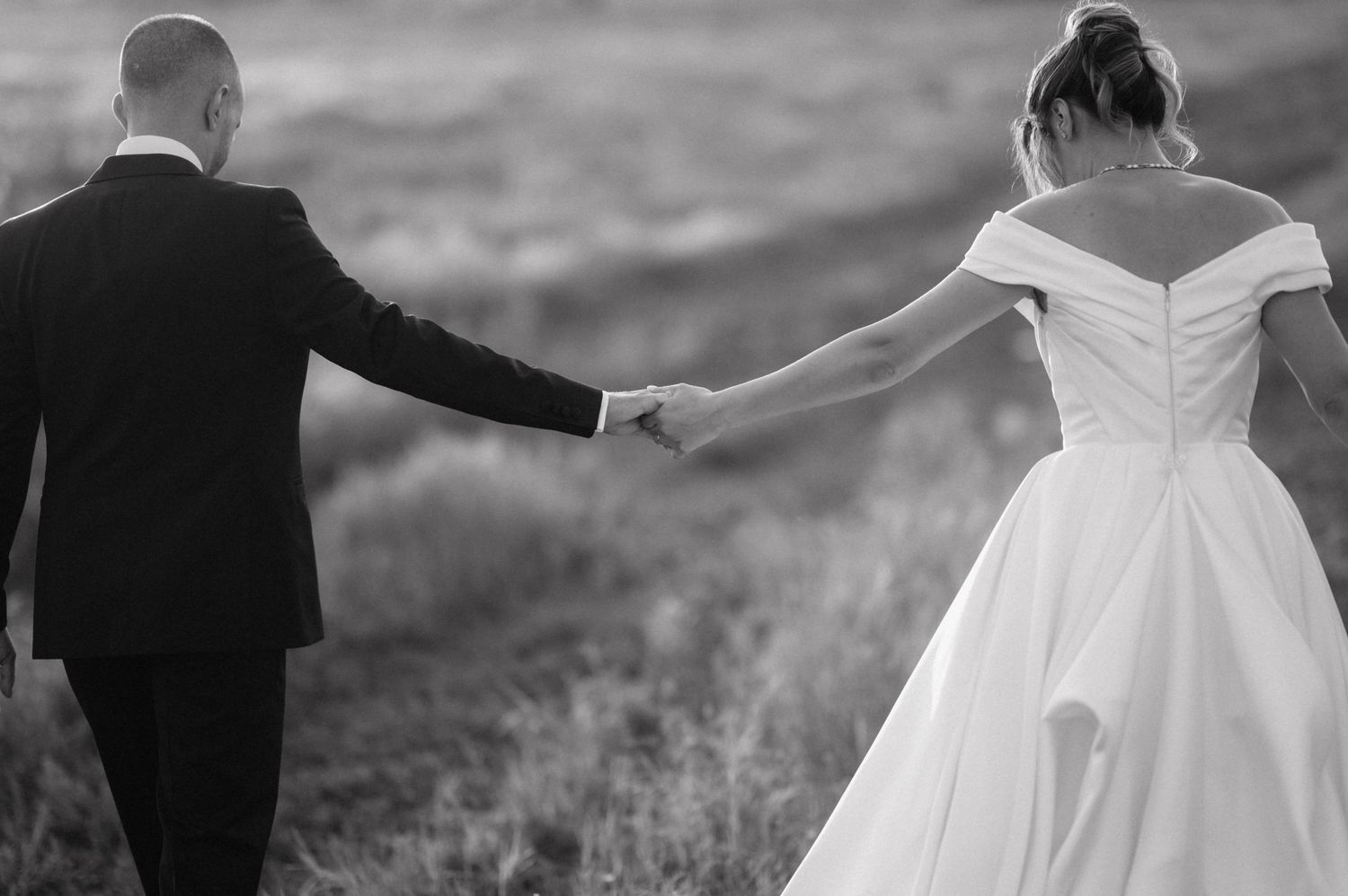 Black and white photo showing a bride in an off-shoulder gown holding hands with groom in suit outdoors.