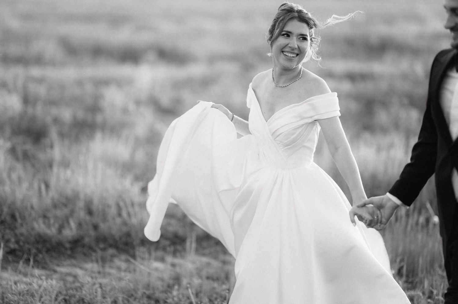 A joyful person in a flowing white wedding dress runs across a grassy meadow in black and white.