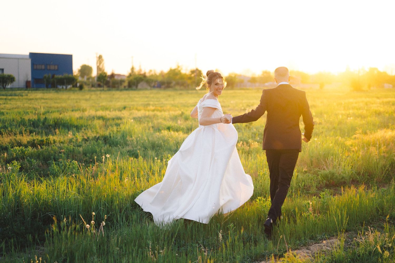 Newlyweds walk hand in hand through a sunlit field at golden hour.