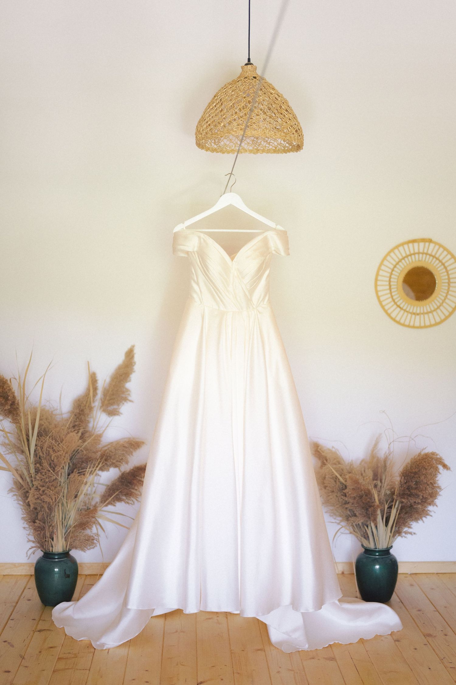 Elegant white wedding dress hanging on display with pampas grass in teal vases and rattan pendant lamp above.