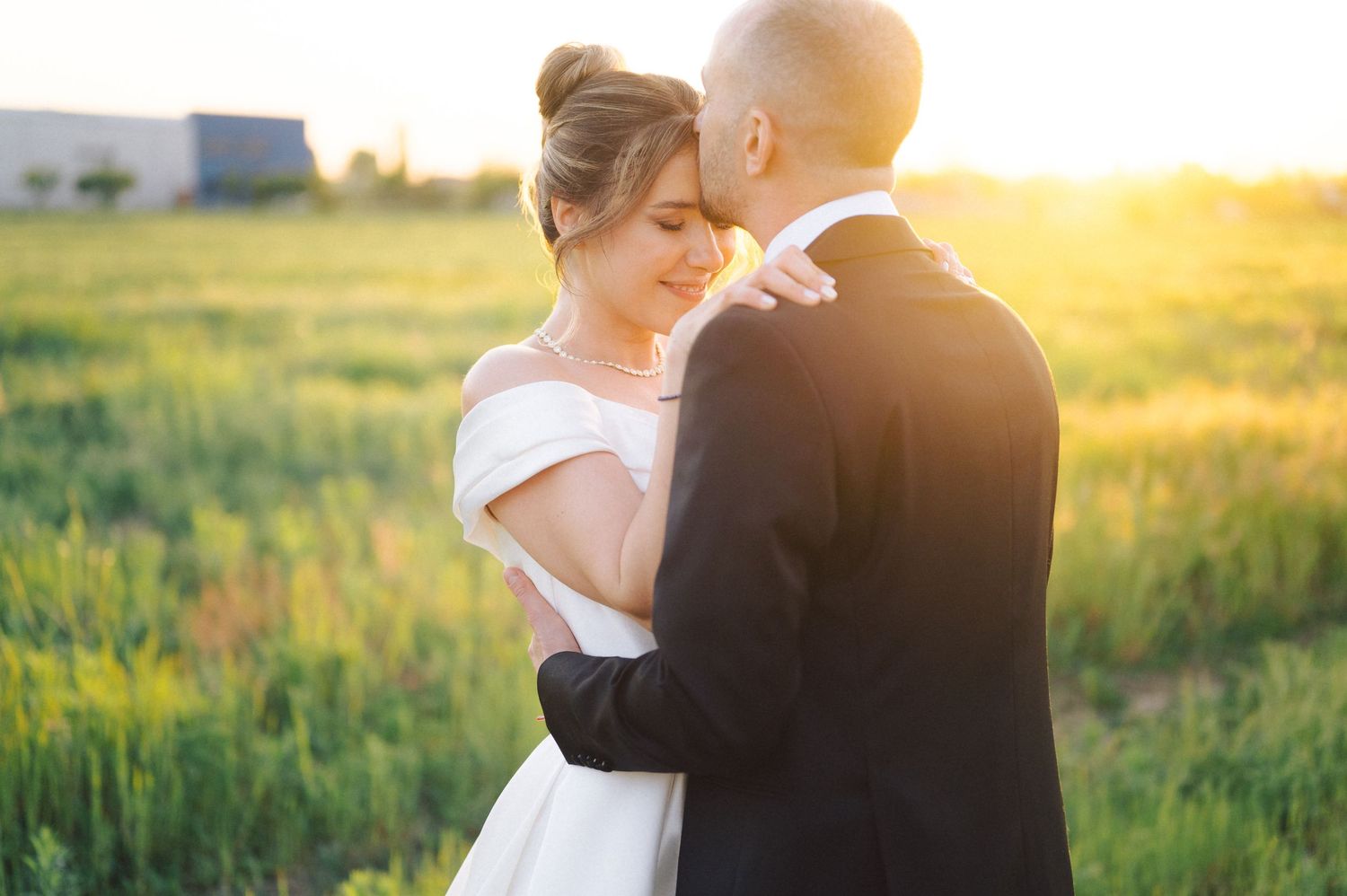 A couple in wedding attire embrace in a sunlit field during golden hour as sunlight glows behind them.