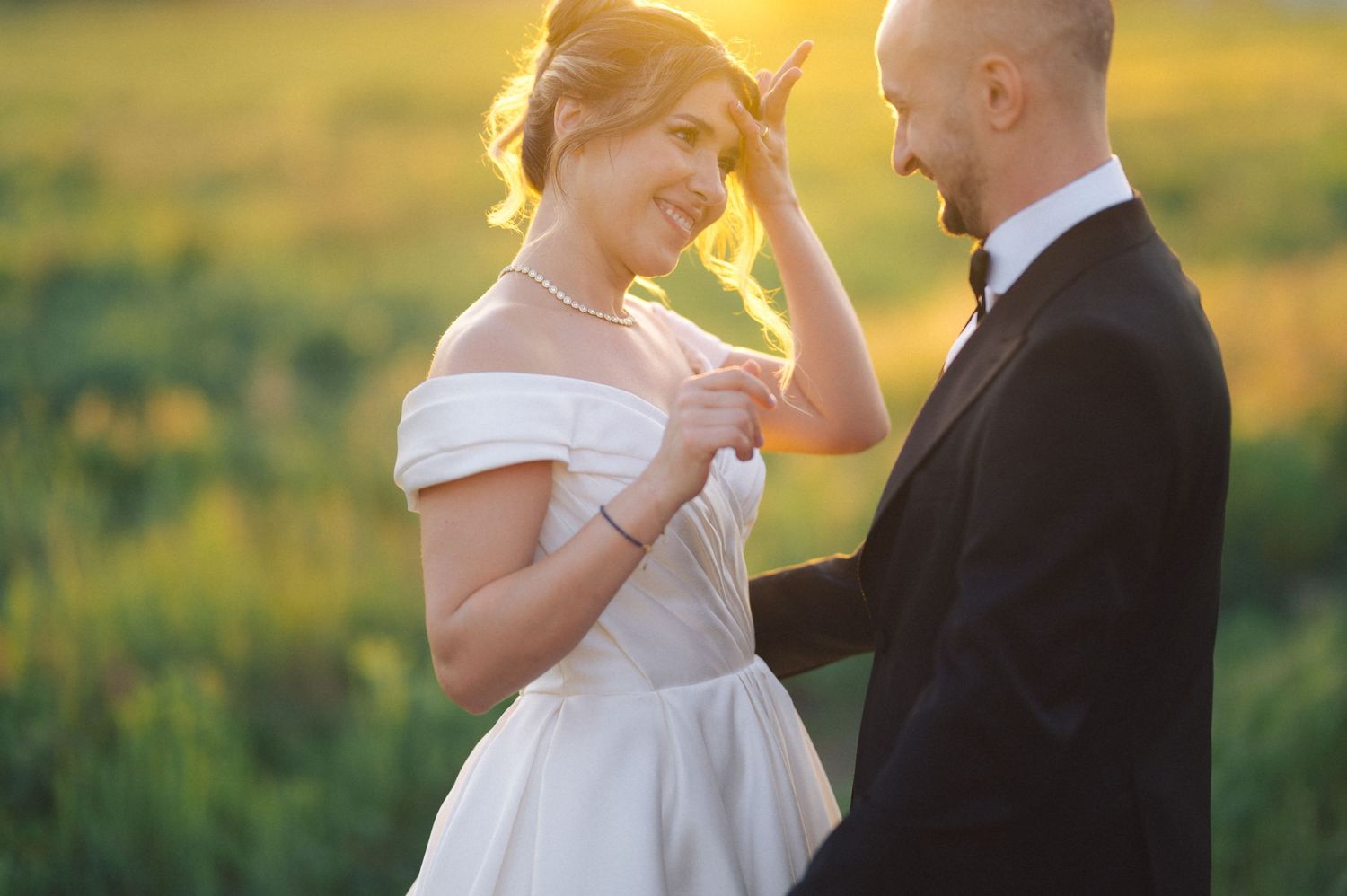 A romantic wedding moment between a couple in a field at golden hour with the bride in an off-shoulder white gown.