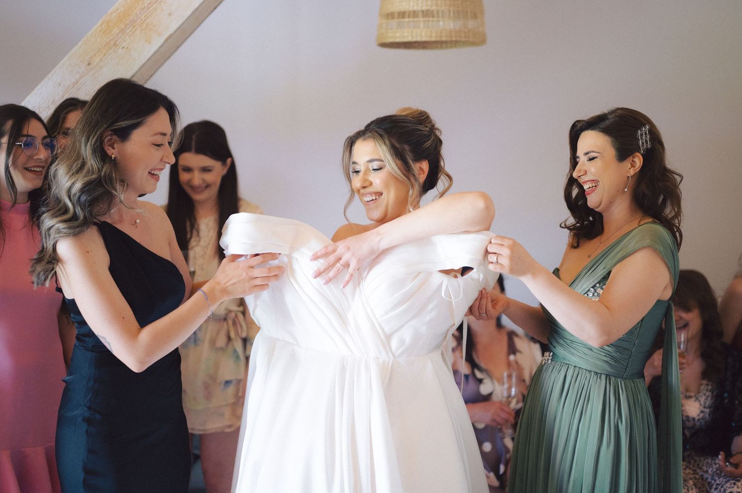 Bridesmaids help dress their companion in an elegant white gown during wedding preparations.